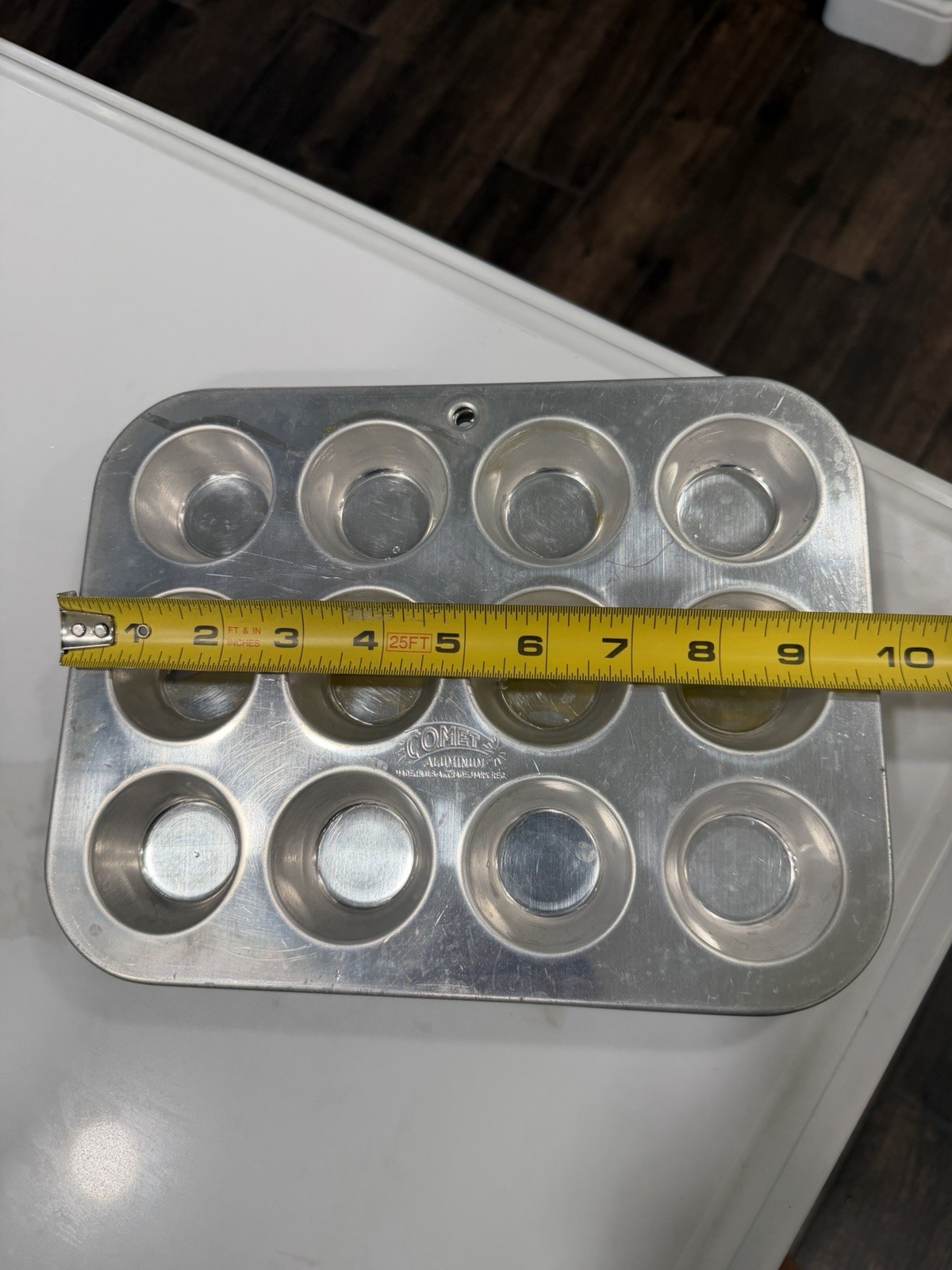 Silver 12-cup mini muffin pan with black circular muffin cups, shown on white surface beside yellow measuring tape.