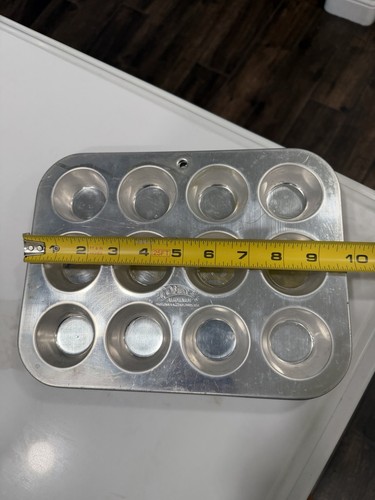 Silver 12-cup mini muffin pan with black circular muffin cups, shown on white surface beside yellow measuring tape.