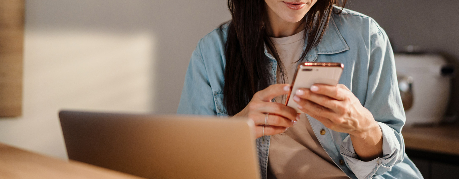 Eine Frau sitzt vor einem aufgeklappten Laptop und blickt auf das Smartphone in ihren Händen.