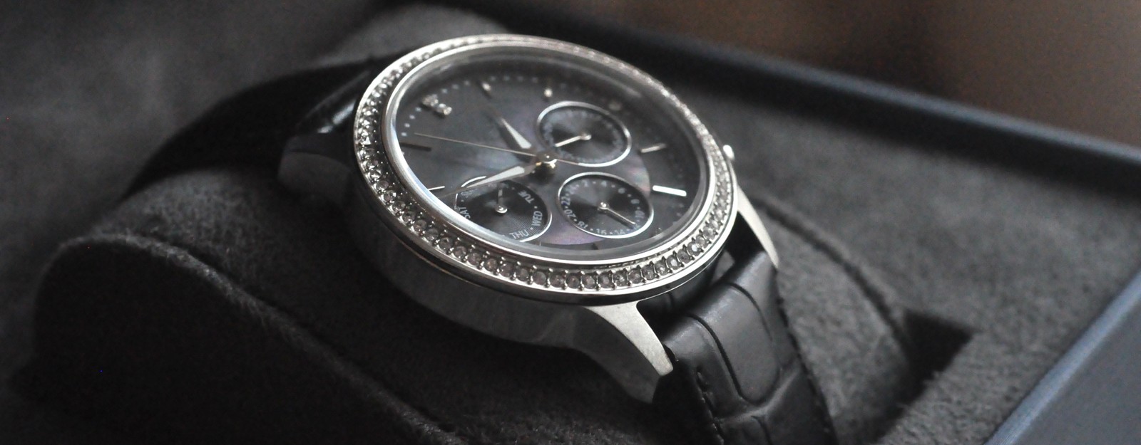 Eine silberne Armbanduhr mit schwarzem Zifferblatt, schwarzem Lederarmband und einer mit Kristallen besetzten Lünette wird in einer schwarzen Uhrenbox präsentiert.