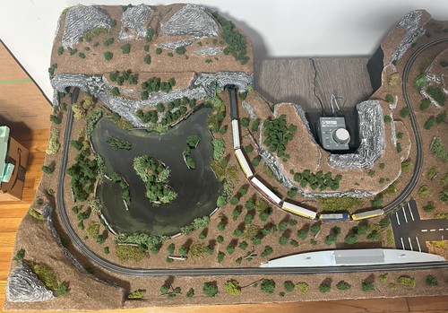 Z-scale model train layout with curved tracks, rocky cliffs, pond, trees, and control panel.