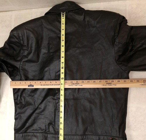 Black leather trench coat with wide shoulder pads, medium size, shown hanging on a wooden ruler against a beige wall.