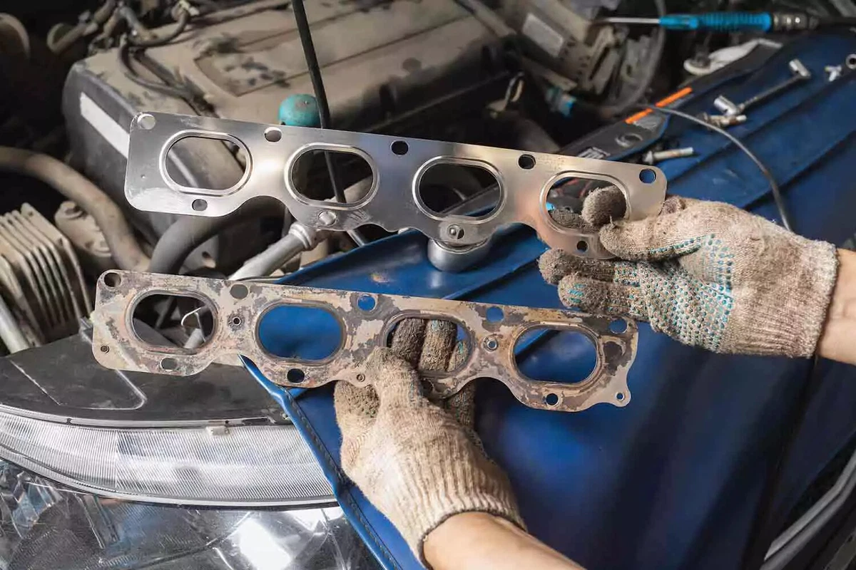 A mechanic wearing white knit work gloves holds a new steel exhaust manifold gasket in his right hand and an old steel gasket in his left hand.