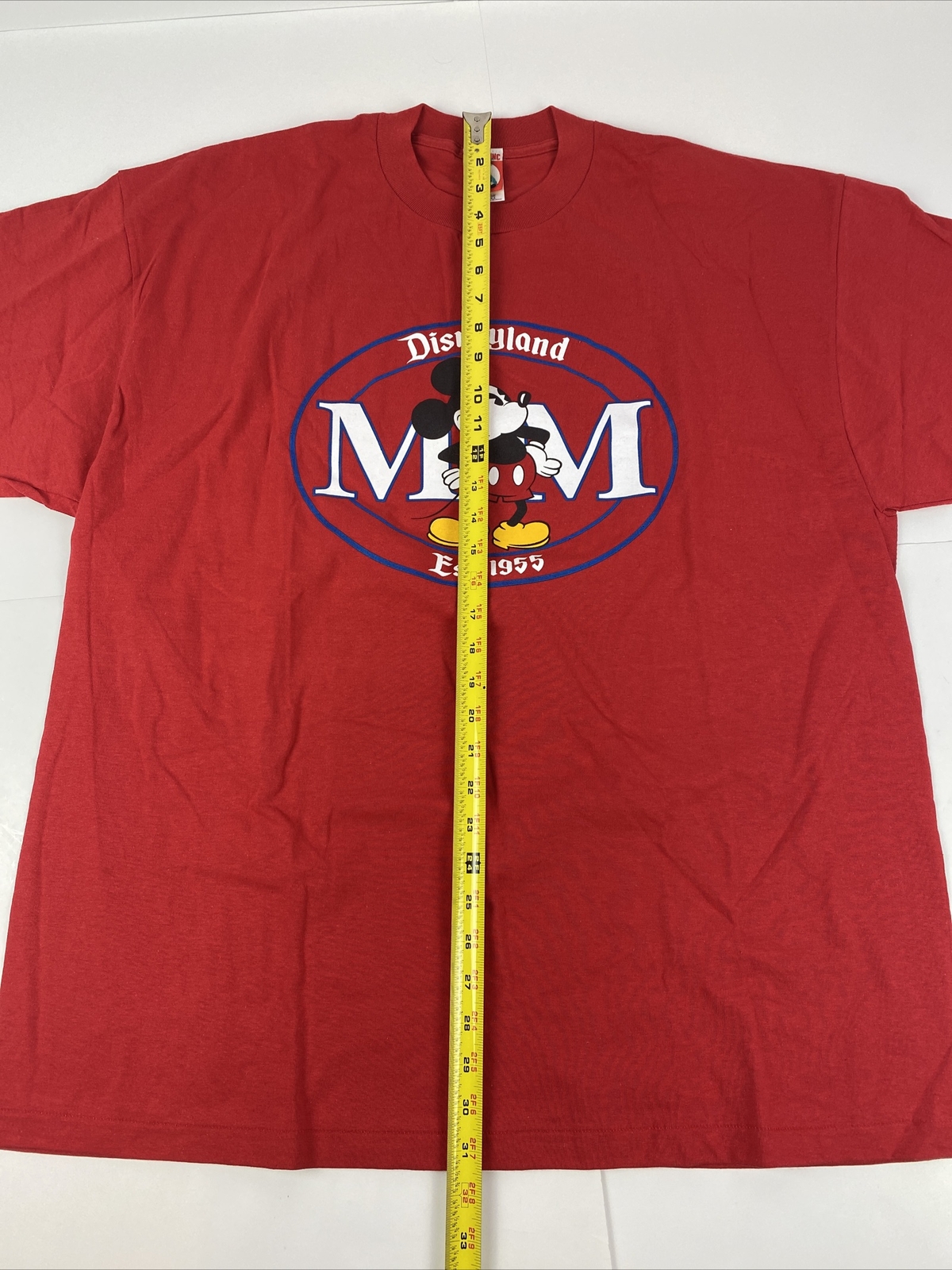 Red vintage Disneyland T-shirt with blue Mickey Mouse logo and “Disneyland Est. 1955” text, shown flat with yellow measuring tape.