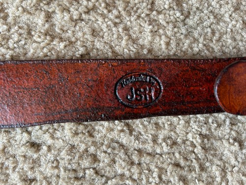 Brown leather leather sling hand with black engraved oval Smith Wesson logo and “JSK” text on tan fabric background.
