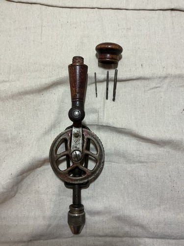Vintage hand crank drill with worn wooden handle, two metal drill bits, and a round wood storage handle on fabric background.