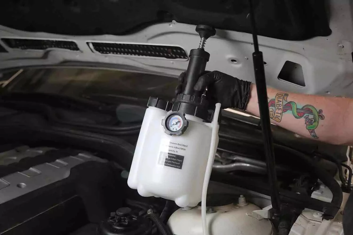 A mechanic wearing a black glove holds a brake bleeder under a vehicle's hood.