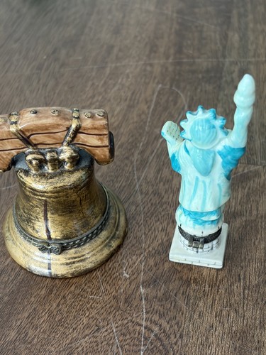 Two miniature Liberty Bell statues, one gold with leather straps, one blue with torch, on a wooden surface.