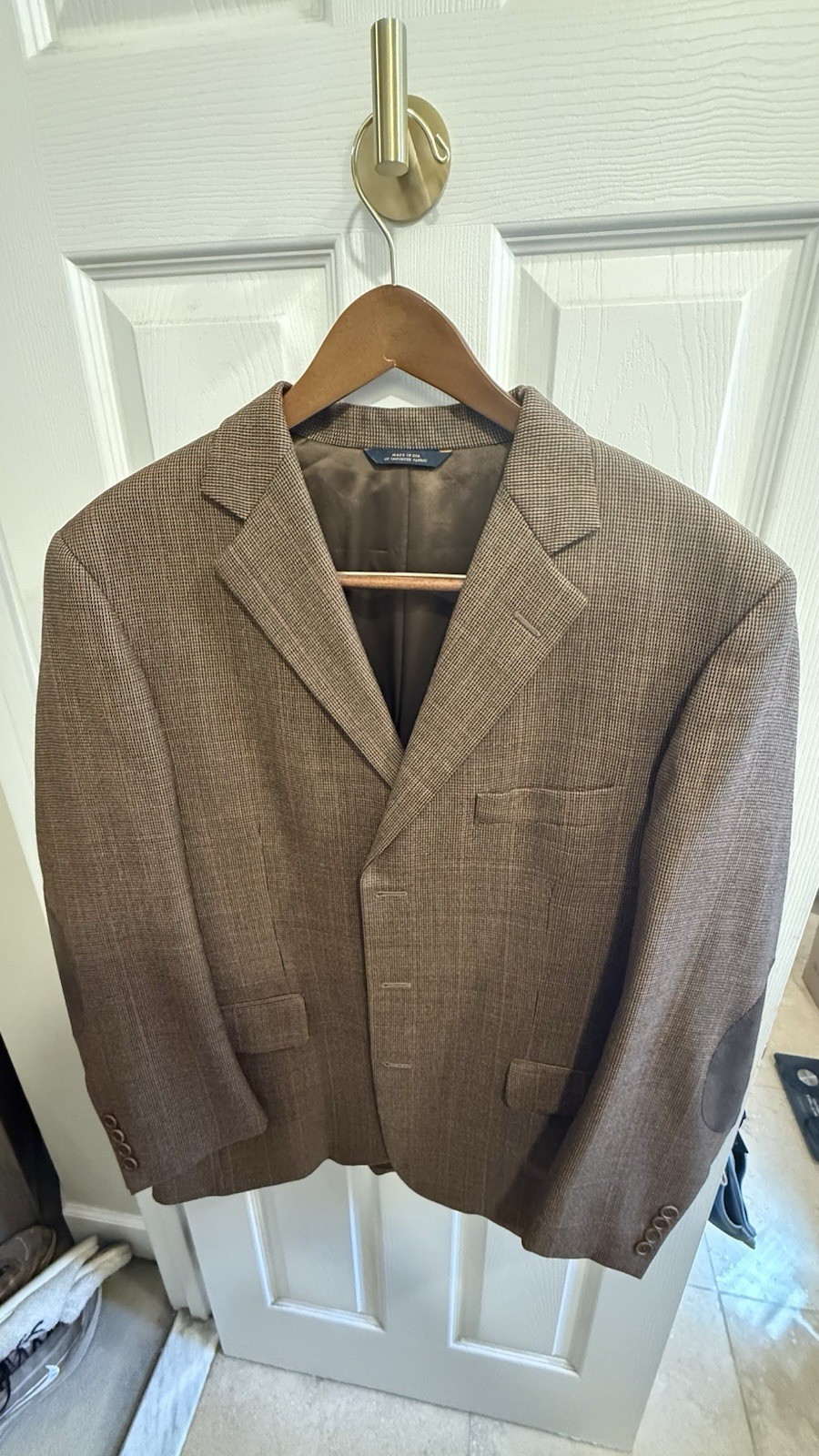 Brown checked tweed blazer with dark leather elbow patches, hanging on a door hook.