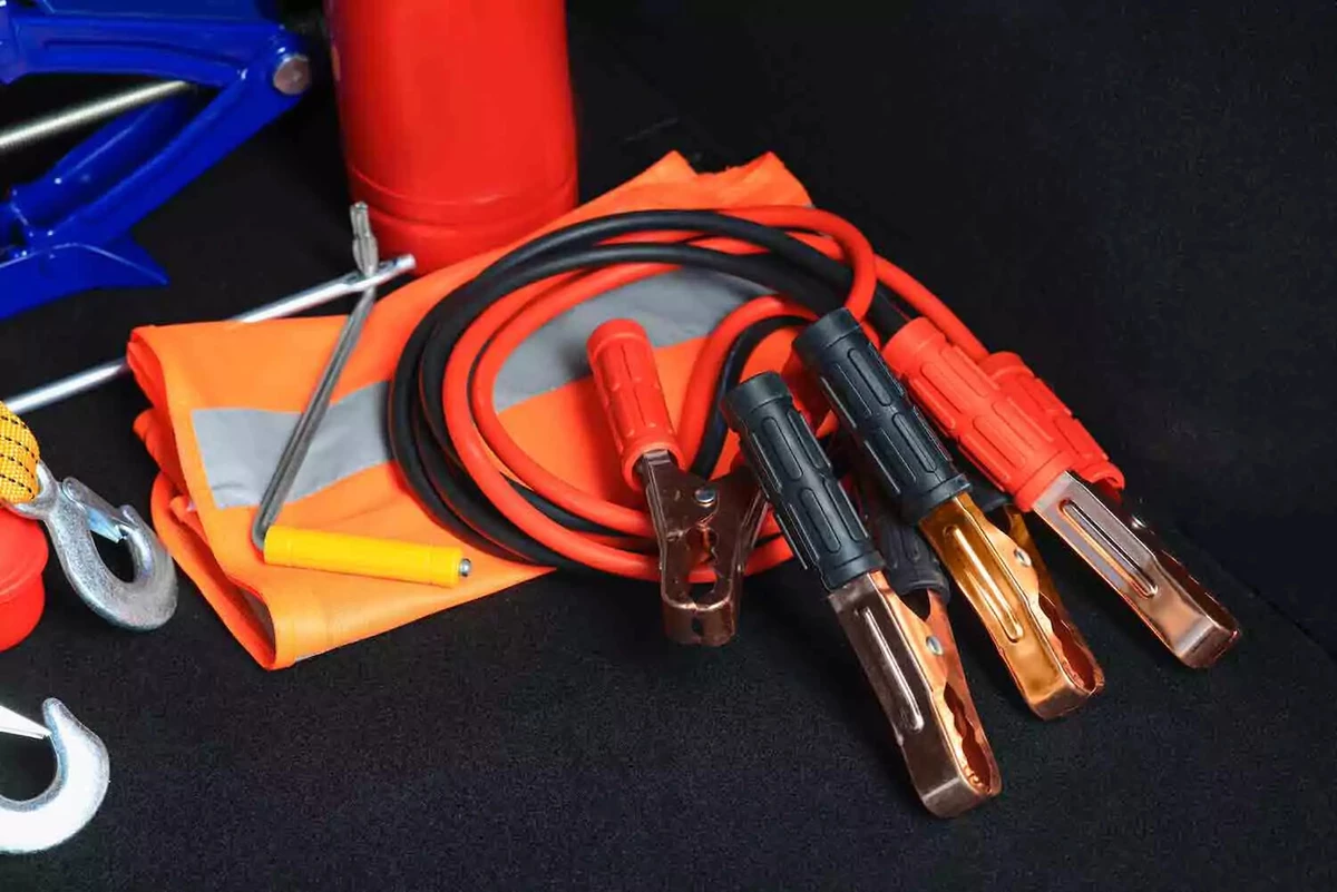 A set of jumper cables in a car trunk, along with a blue scissors jack, a red fire extinguisher, an orange safety vest, and a tow rope.