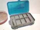Open teal 20-compartment fishing vest with yellow logo, surrounded by three assorted vintage lures in a clear Plano Mini Mag case.