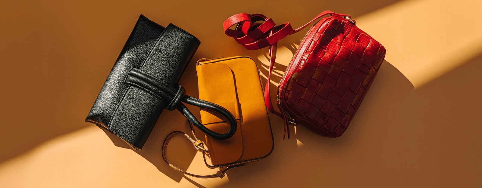 Drei stilvolle Handtaschen in Schwarz, Gelb und Rot, angeordnet auf einem warmen orangefarbenen Hintergrund, präsentieren ihre verschiedenen Designs und Texturen.