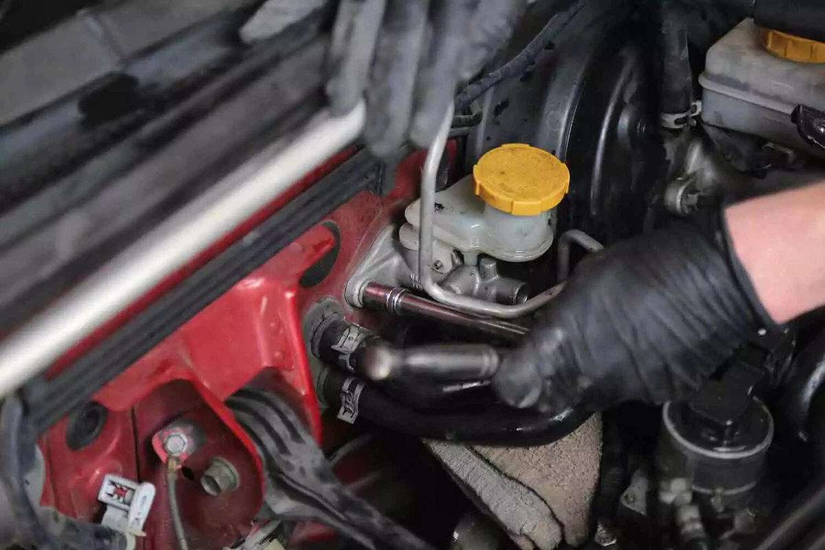 A mechanic wearing black gloves uses a socket wrench to unbolt the clutch master from the firewall.