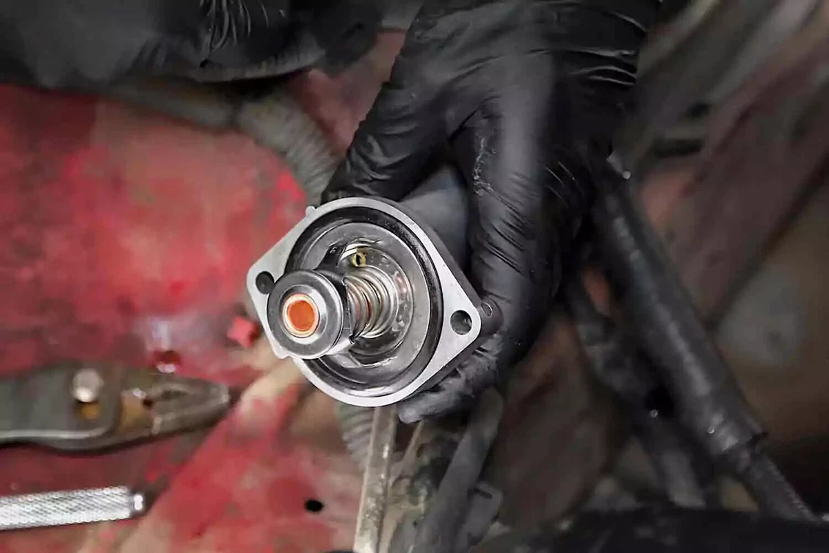 A mechanic wearing a black nitrile glove holding a thermostat, with a red car’s engine bay and a set of pliers in the background.