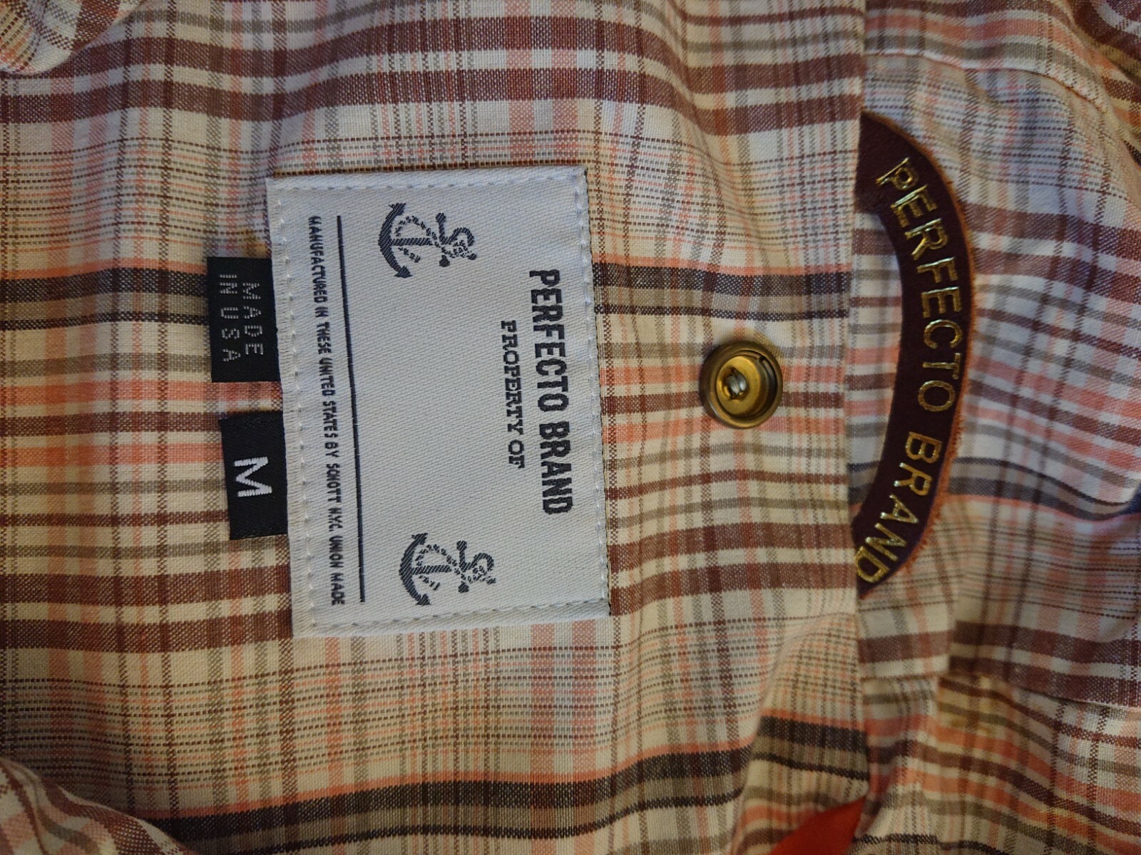 White fabric tag with black anchor logo and “Perfecto Brand” text inside yellow buttonhole on 60/40 cotton nylon parka.