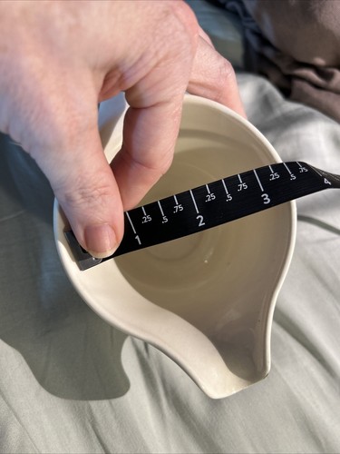 Hand measuring the interior diameter of a white ceramic bowl with black floral print, using a dark diagonal ruler for scale.