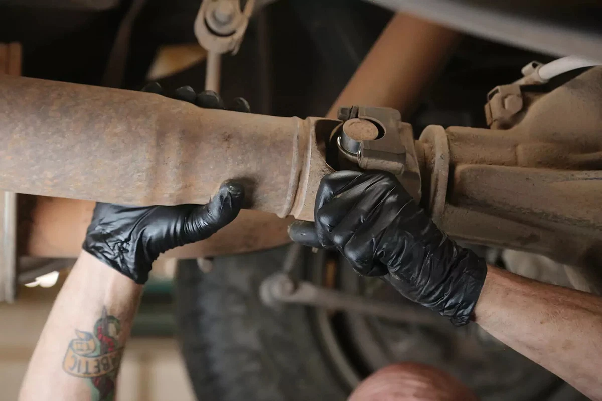 A mechanic wearing black gloves reattaches the driveshaft to the transmission.