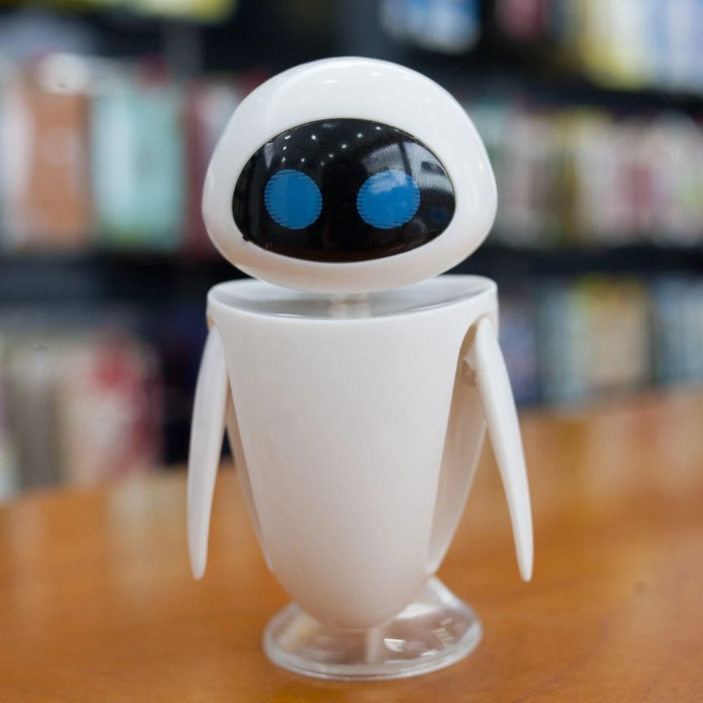 White plastic robot toy with black oval face, blue eyes, and articulated arms on clear stand, set against bookshelf background.