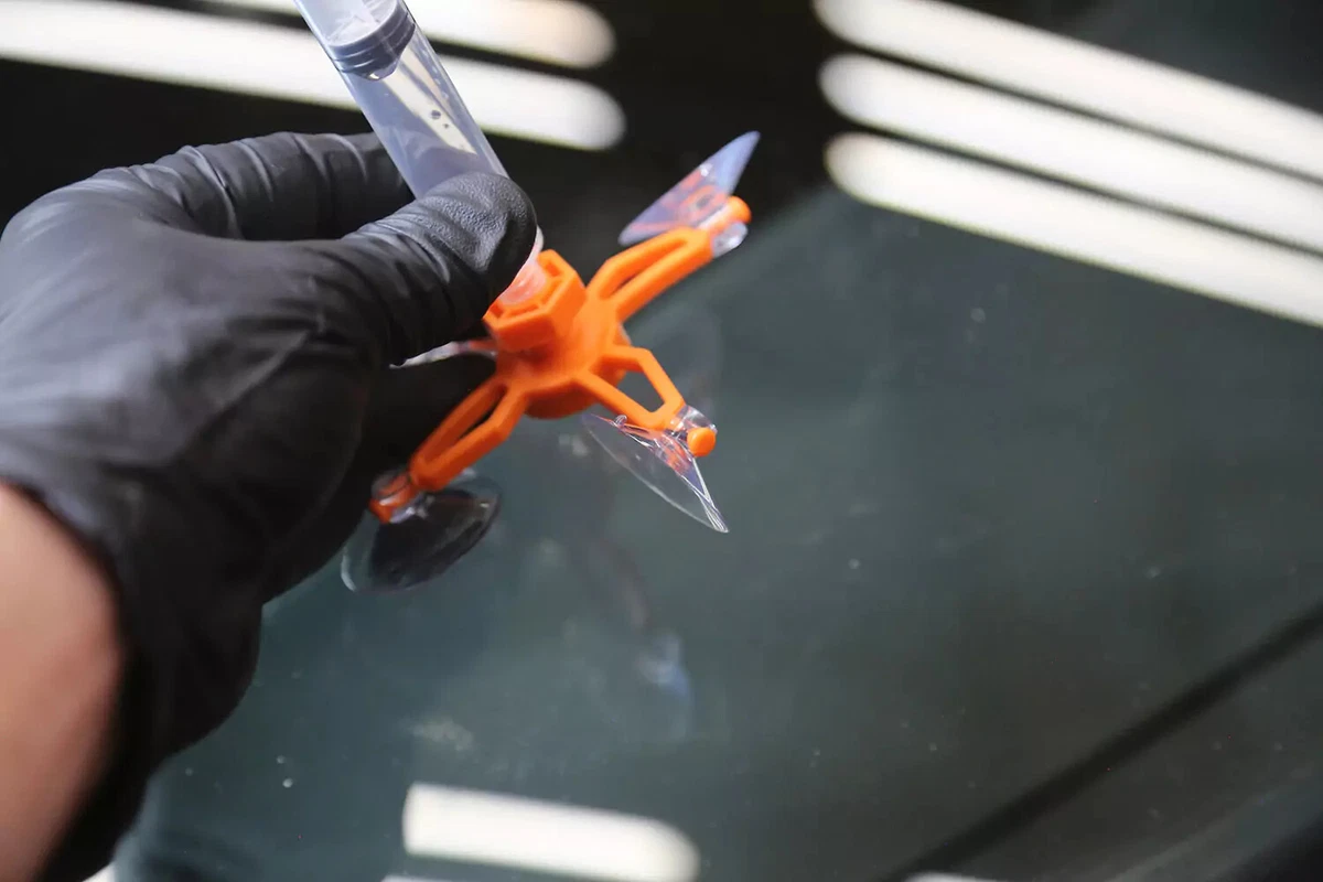 A mechanic wearing a black glove removes the repair jig and syringe from the vehicle's windshield.