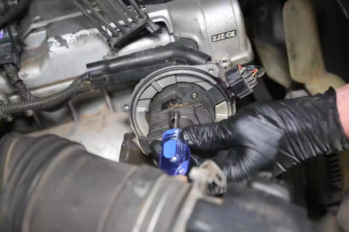 A mechanic wearing black gloves uses a screwdriver to remove the rotor from a vehicle's distributor cap.