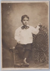 Niño con pajarita grande y bragas en silla de mimbre, c1910 estudio retrato RPPC - Imagen 1 de 2