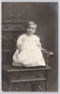 Postal RPPC Infantil Niña en Vestido Blanco Sentado en Silla Grande Adornada 1904-1918 - Imagen 1 de 2