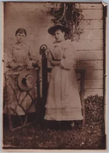 Dos mujeres jóvenes, sombreros y vestidos, al lado de la cerca, años 1900-20, foto sepia - Imagen 1 de 2