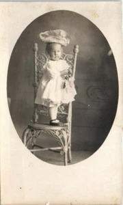 1910er Jahre RPPC Postkarte Kind Baby Mädchen mit riesiger Mütze ? Ausgefallenes Stuhlkleid - Bild 1 von 2