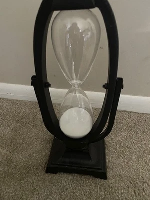 Un moderno reloj de arena decorativo con marco de metal negro y arena blanca Foto 1 de 4