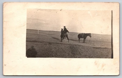 Hombre montando caballo oscuro en campo abierto y caballo ensillado de pie cerca RPPC Foto 1 de 2