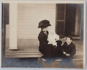 Mujer en sombrero sosteniendo bebé con niño sonriente, escena de porche, foto sepia de 1900 - Imagen 1 de 2