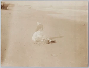 Niña con vestido y arco juega en la playa con cubo, foto sepia de 1913 - Imagen 1 de 2