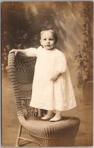 Foto real vintage de los años 1910 STUDIO RPPC postal niña en vestido blanco/silla de mimbre - Imagen 1 de 2