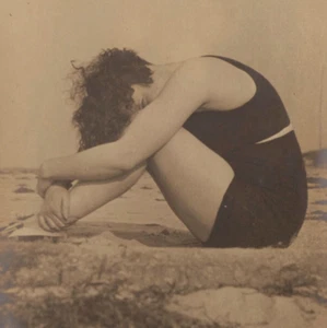 Foto antigua de una mujer tímida escondiendo rostro posando en arena en traje de baño negro - Imagen 1 de 2